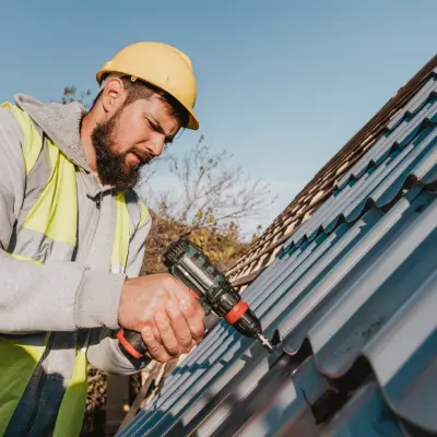 A construction worker in a yellow hard hat and reflective vest uses a power drill on a metal roof under a clear blue sky A construction worker in a yellow hard hat and reflective vest uses a power drill on a metal roof under a clear blue sky